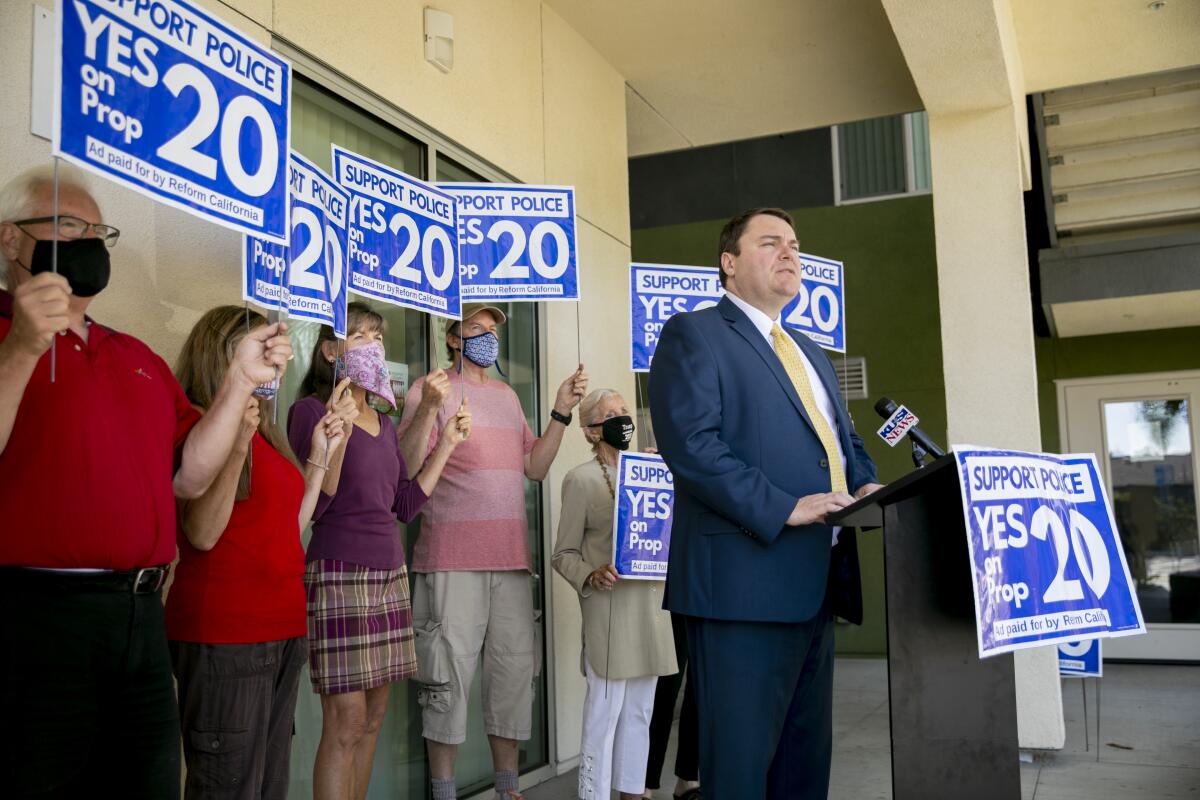 People holding Yes on Prop 20 signs stand behind a man speaking at a lectern during a news conference