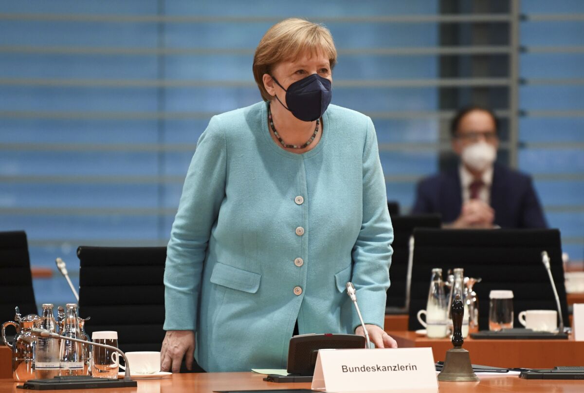 A woman in a pale blue jacket and dark mask stand before a microphone and sign that reads Bundeskanzlerin
