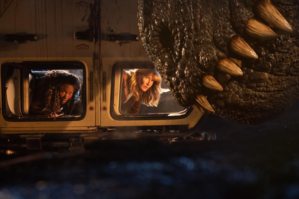 Two women inside an upside-down vehicle look out the windows and see the teeth of a huge dinosaur
