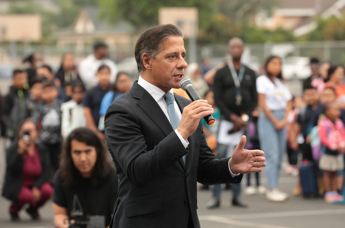 Alberto Carvalho, in suit and tie, speaks into a mic in a school playground, with children and adults behind him.