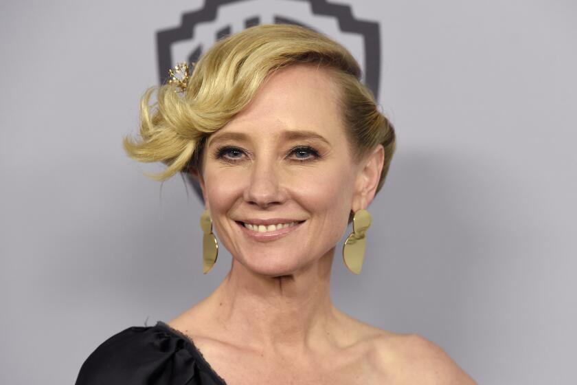 A woman with blond hair in an updo posing in gold earrings and an off-shoulder black dress