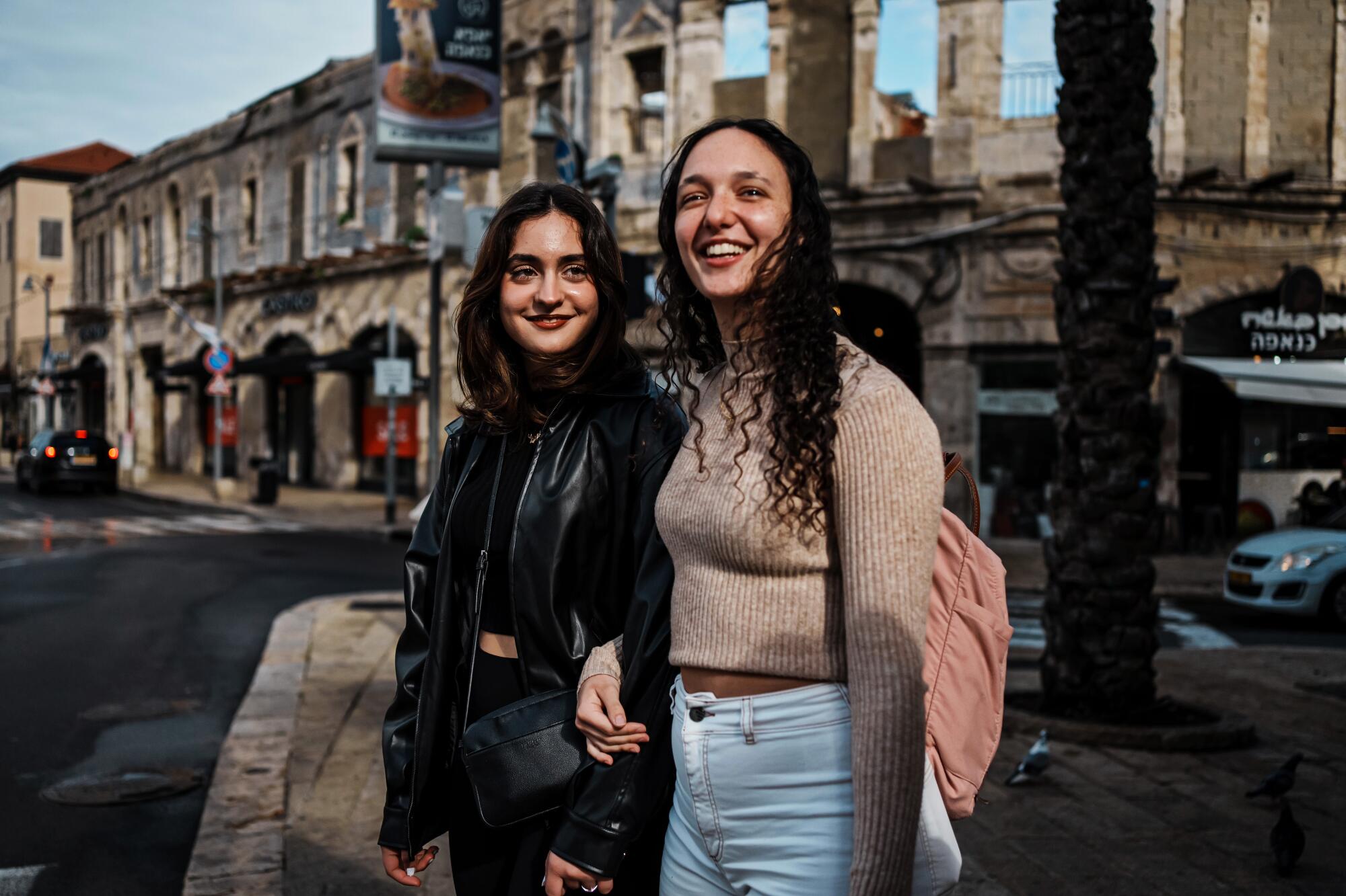 Two teen girls stand close together smiling on a street 
