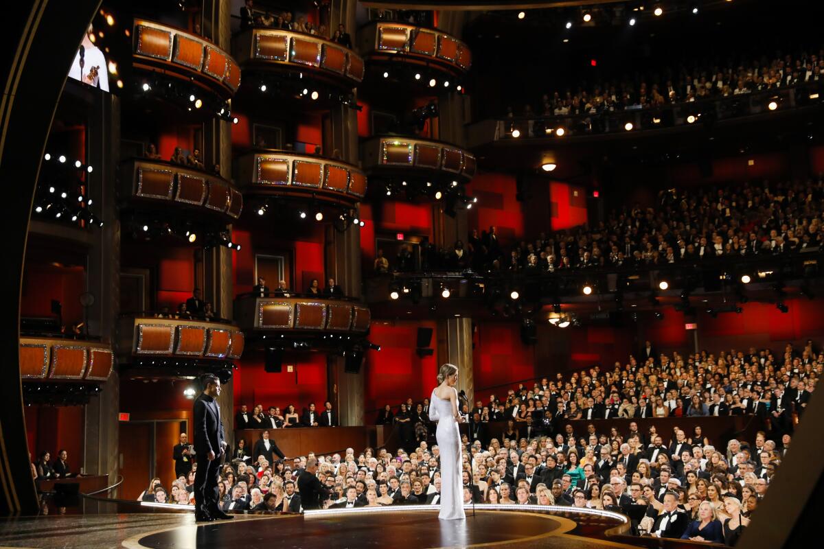 Renee Zellweger stands on the Oscars stage in a white gown before a packed theater