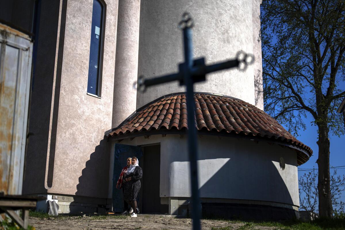 Worshippers walk out a small door in a side building of a church. A cross is in the foreground.