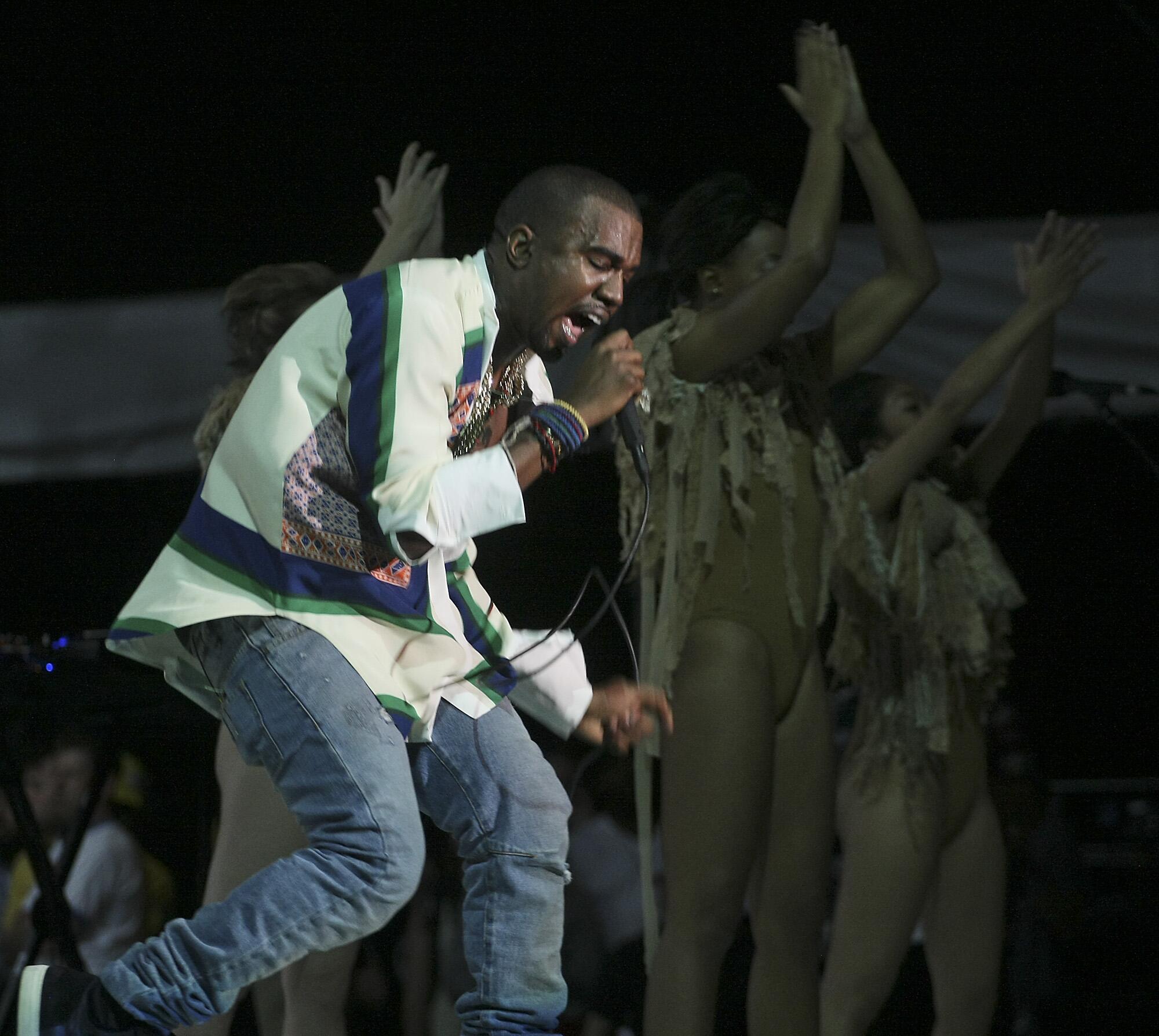Kanye West raps into a microphone with dancers down  him