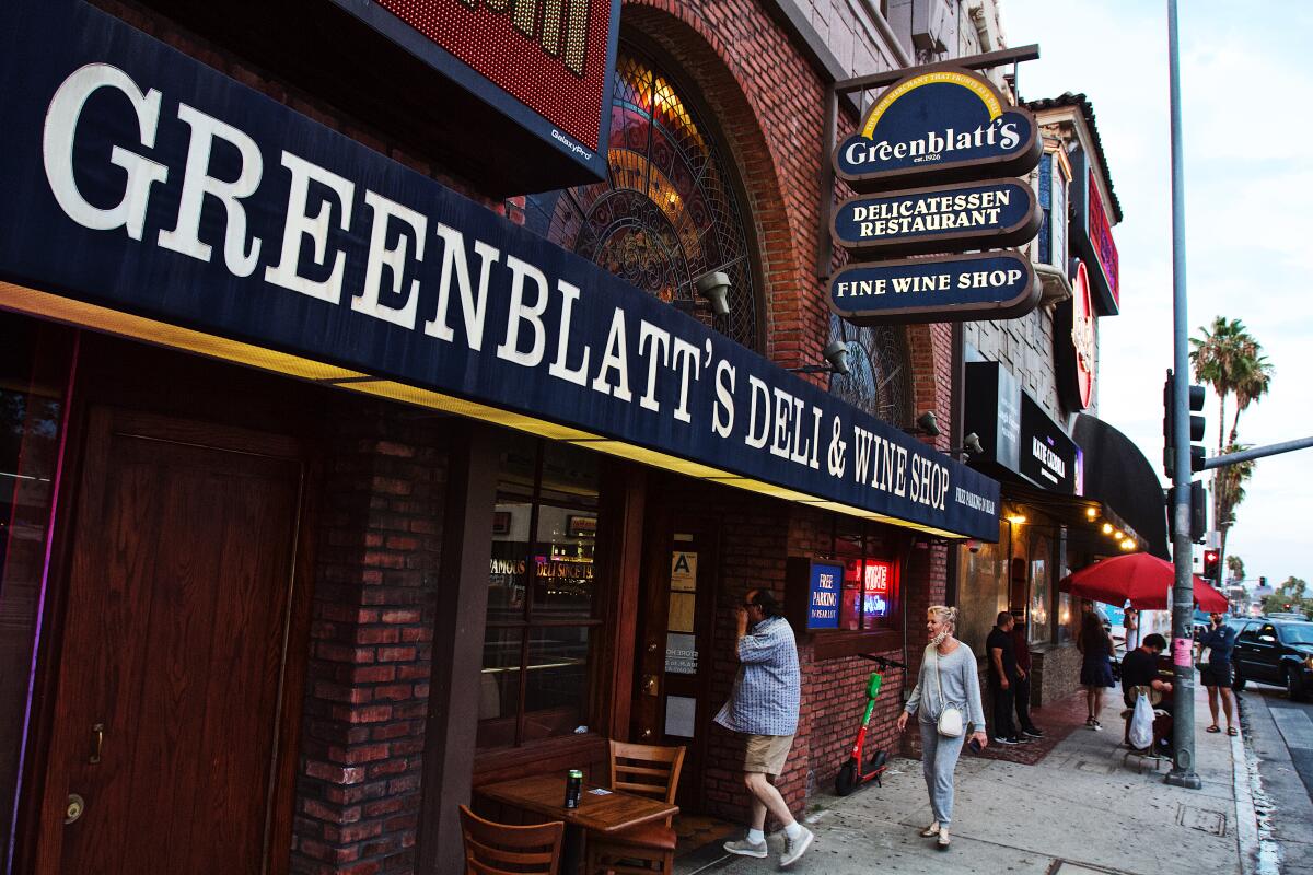 A sign saying "Greenblatt's" hangs over another sign as people walk from the street into the front of a shop.