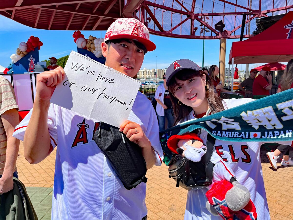 Two Shohei Ohtani fans from Japan wear Angels gear. A man holds a white piece of paper with a message on it.