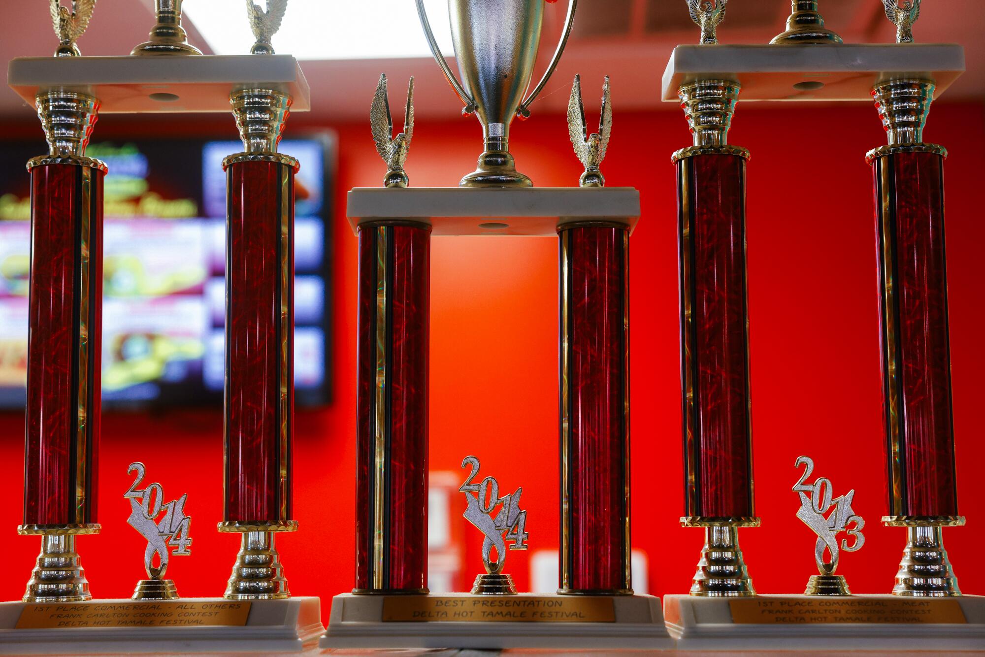 A row of trophies in front of a red wall at a tamale restaurant.