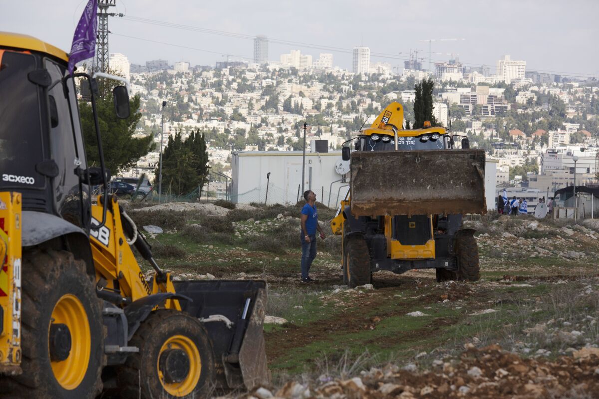 A man talks to the driver of a bulldozer in a rocky field against the backdrop of a city.