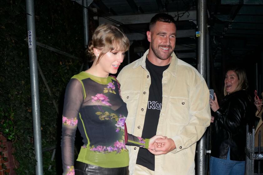 Taylor Swift in a sheer top and black skirt holding hands with Travis Kelce in a beige jacket-pant set and black shirt