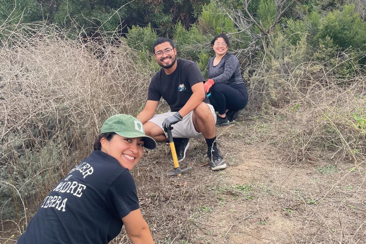 Three people smile at the camera while weeding at a restoration event. 