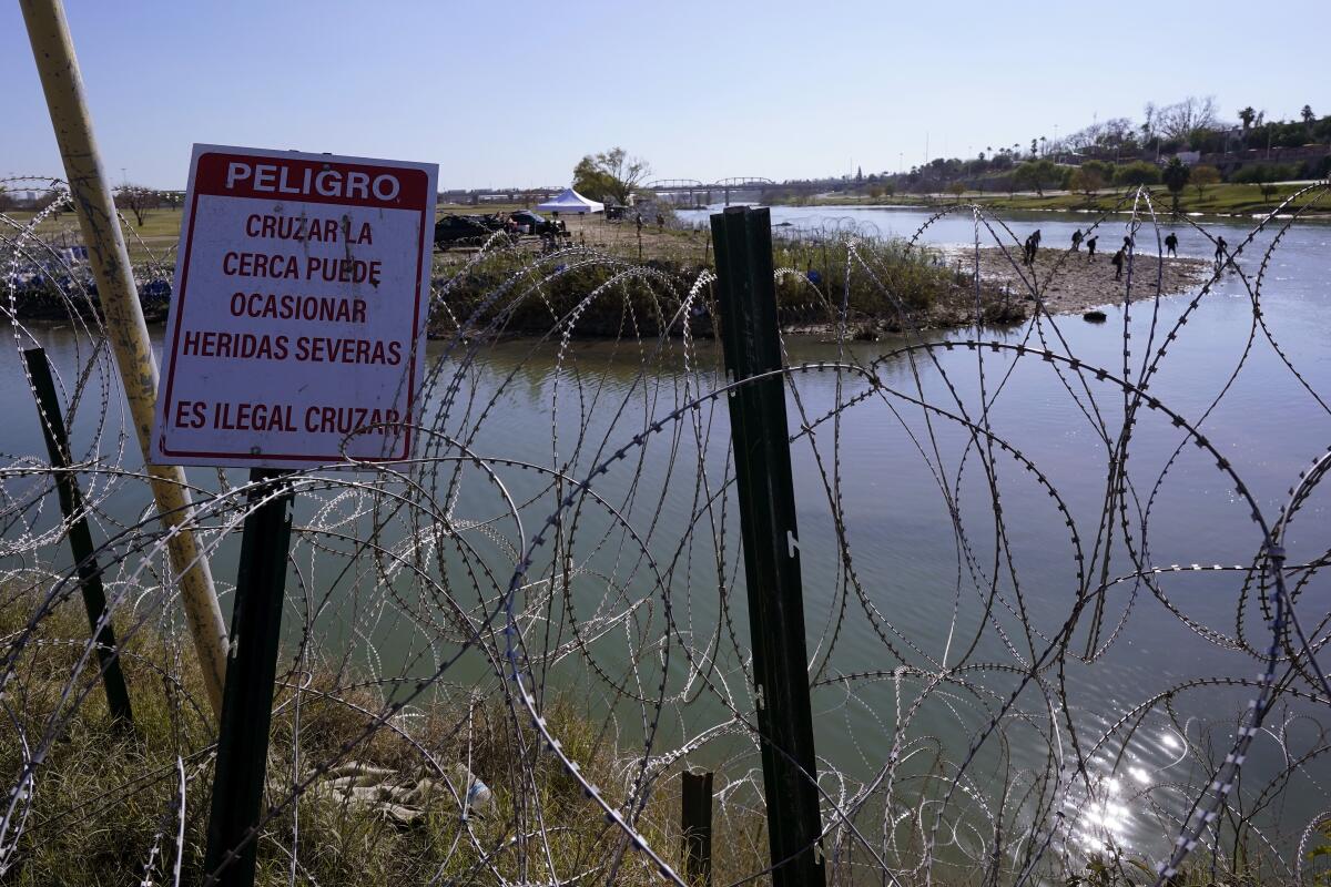 Migrants cross the Rio Grande into the U.S. behind barbed wire and a sign saying it's dangerous and illegal to cross.