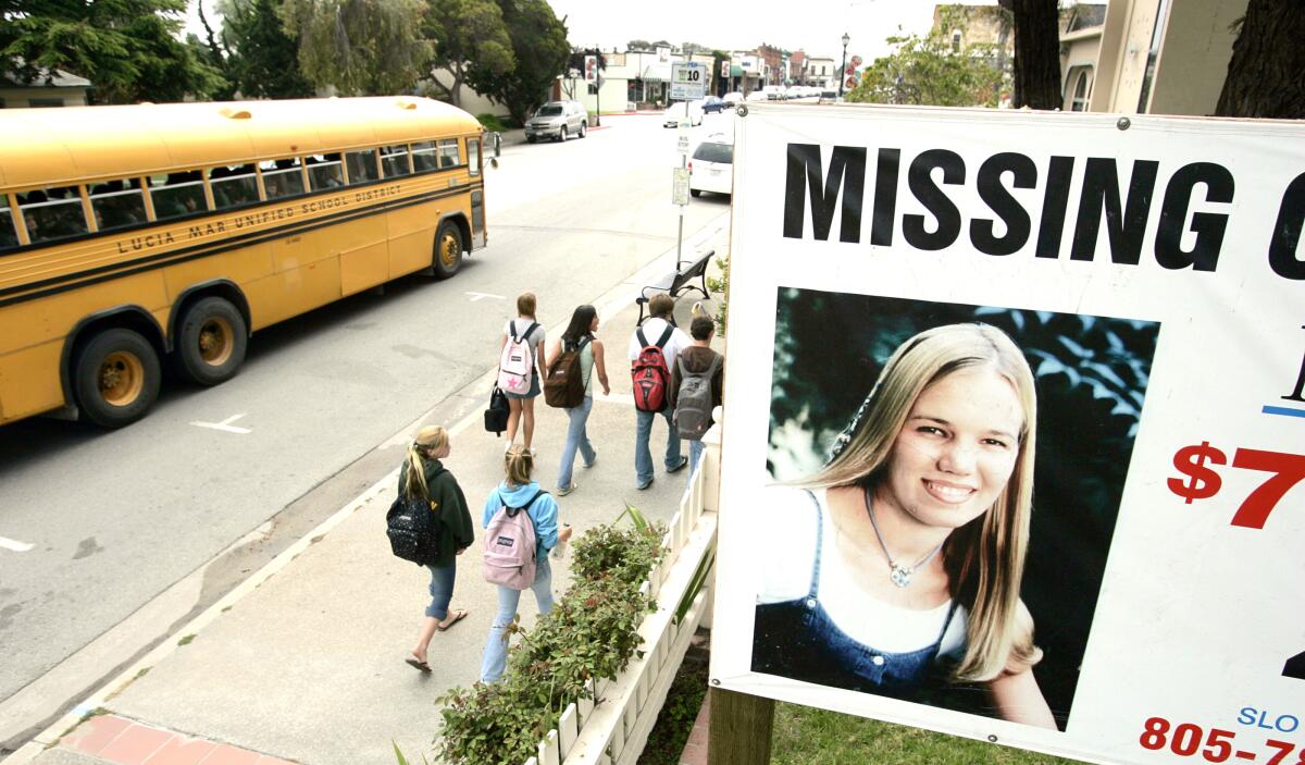 Several students walking on a sidewalk below a poster of a missing young woman as a school bus drives by