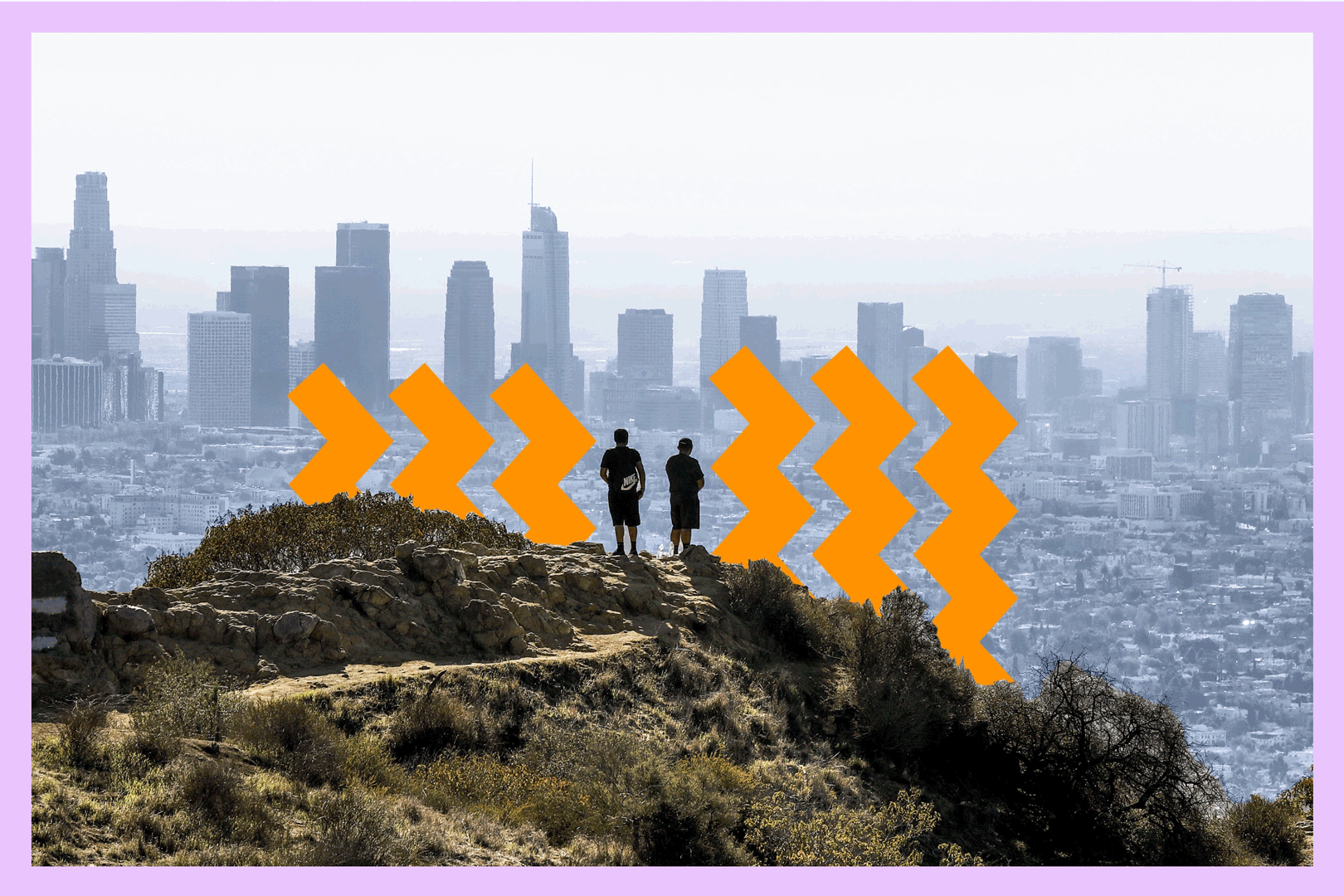 An aerial view of two hikers on a hillside with the cityscape of Los Angeles behind them