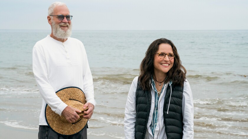 A bearded man holding a hat and a women stand on a beach.