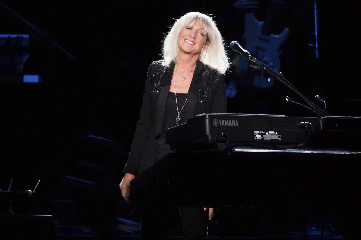 A woman with white-blond hair in a sparkly black blazer stands onstage next to a piano
