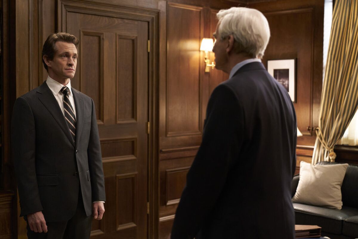 A younger man and an older man speak in a wood-paneled office