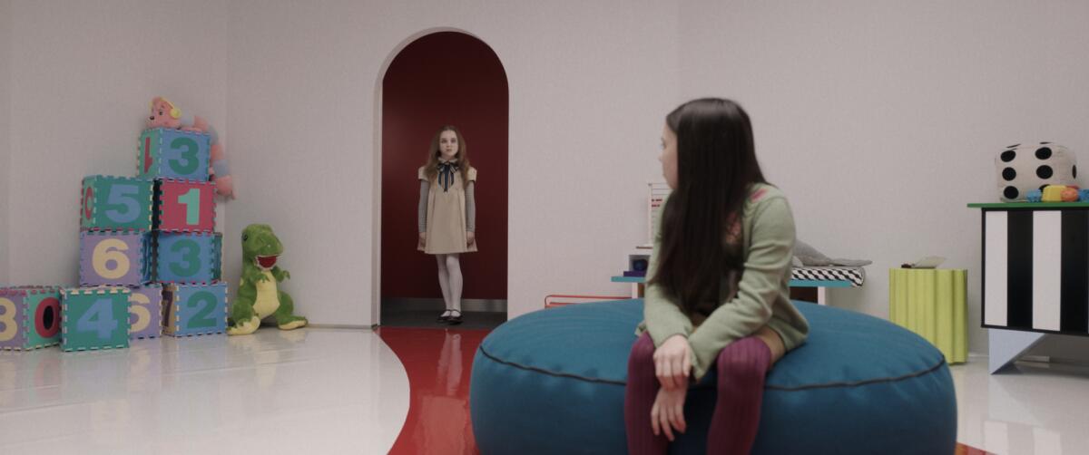 A young girl sits on a round pouf looking over her shoulder at her robot companion, which looks like a young girl