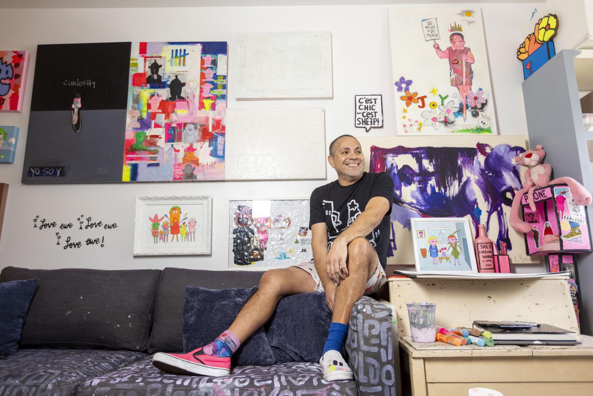 A man sitting on the edge of a couch with art covering his wall. 