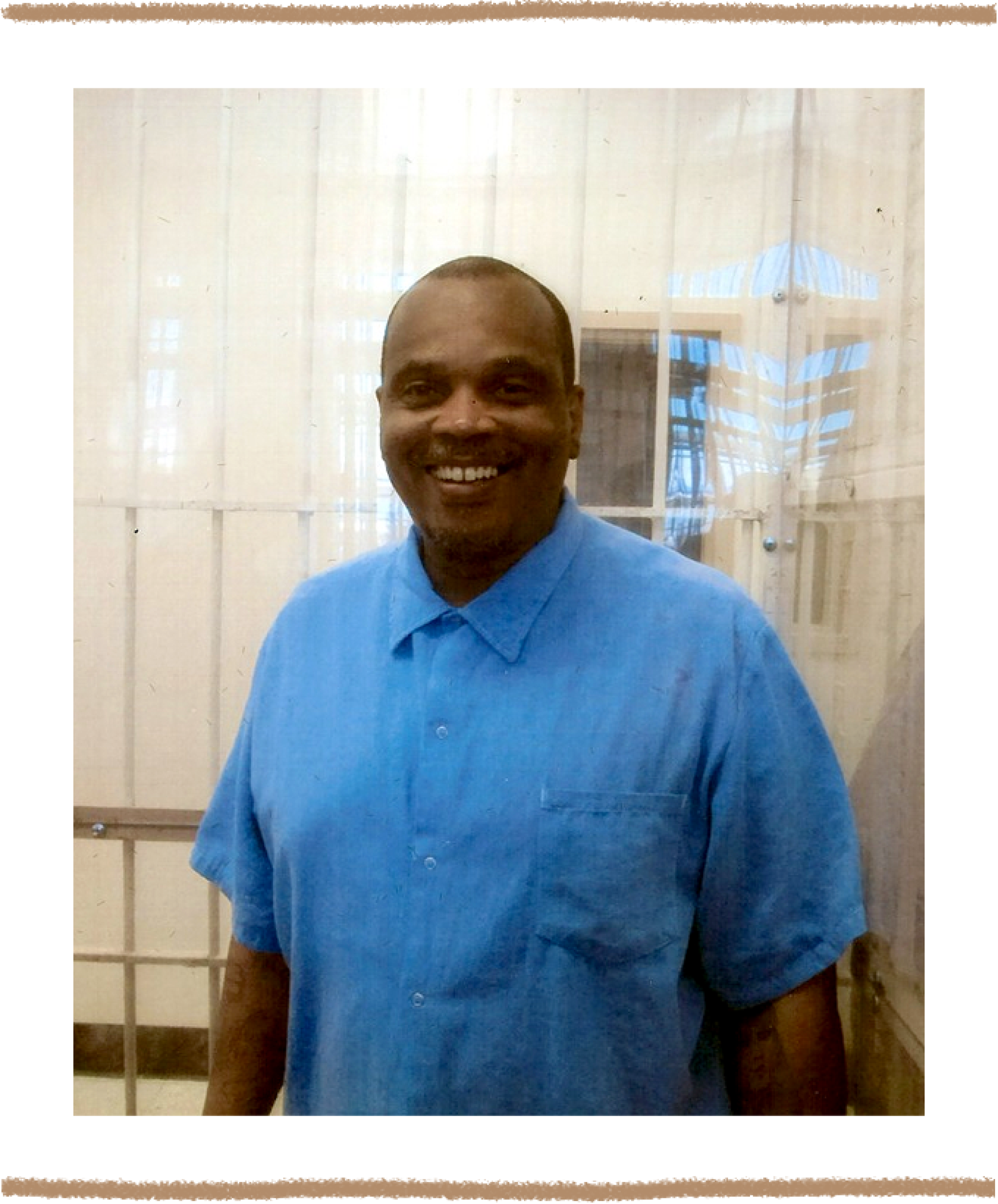 A photo of a man in a blue button-down shirt smiling for a photo in front of white bars