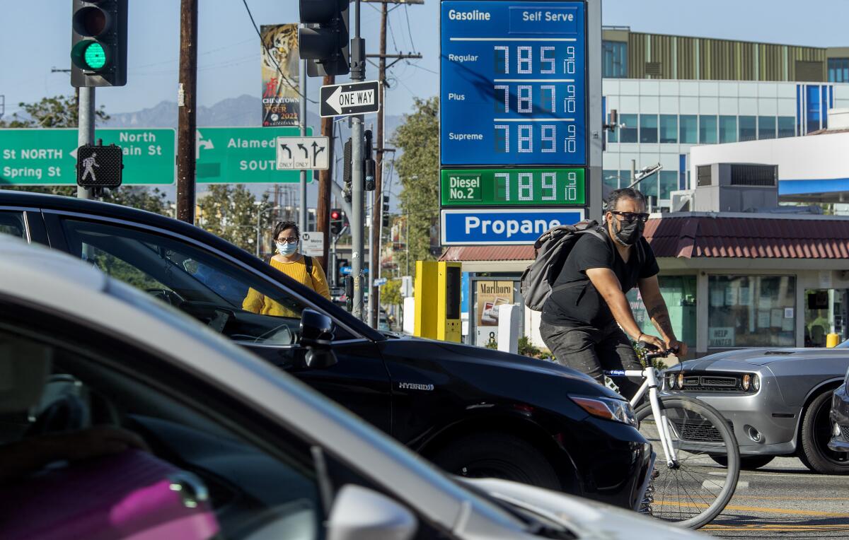 A man on a bicycle crosses a street between cars in front of a gas station sign with prices from $7.85 to $7.89 per gallon