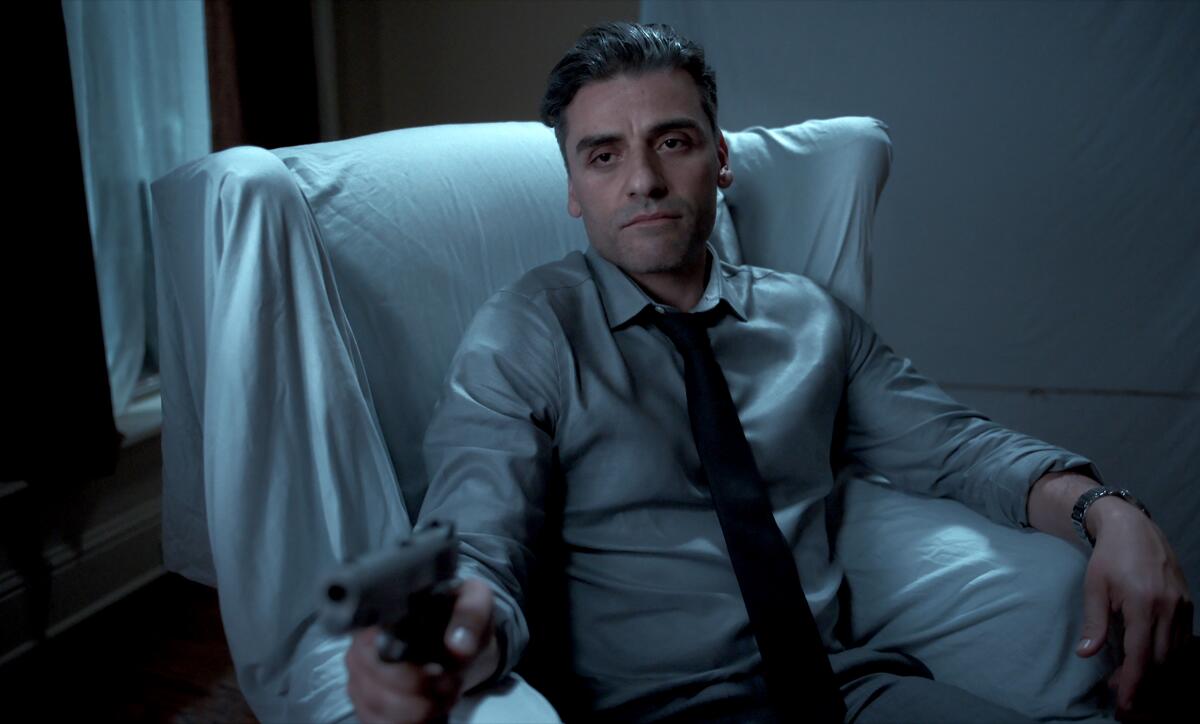A man sits in a sheet-covered chair in a dark room, aiming a pistol toward the camera.