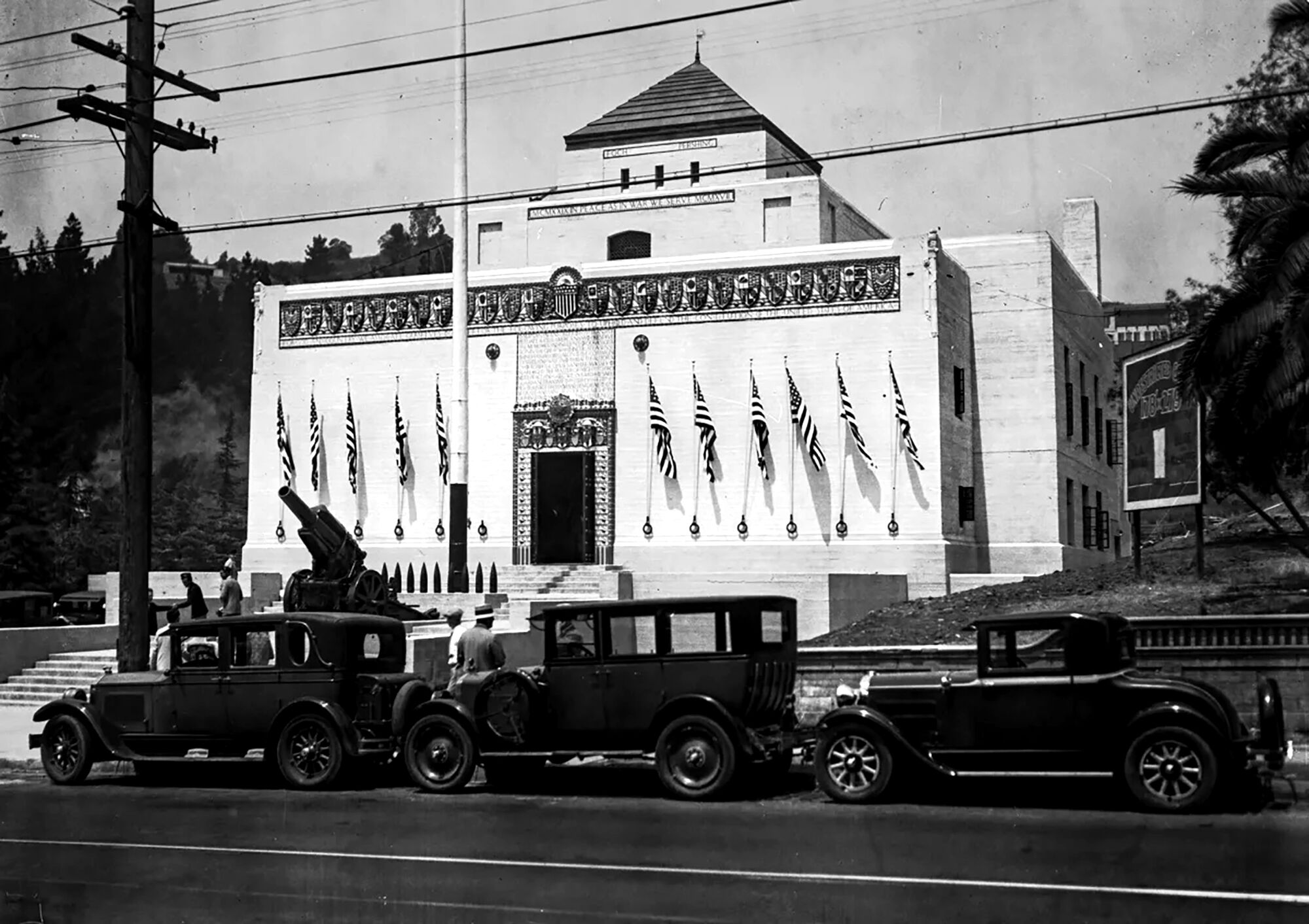 1920s automobiles are lined up in front of an American flag-lined Egyptian Revival-style building, with a cannon in between