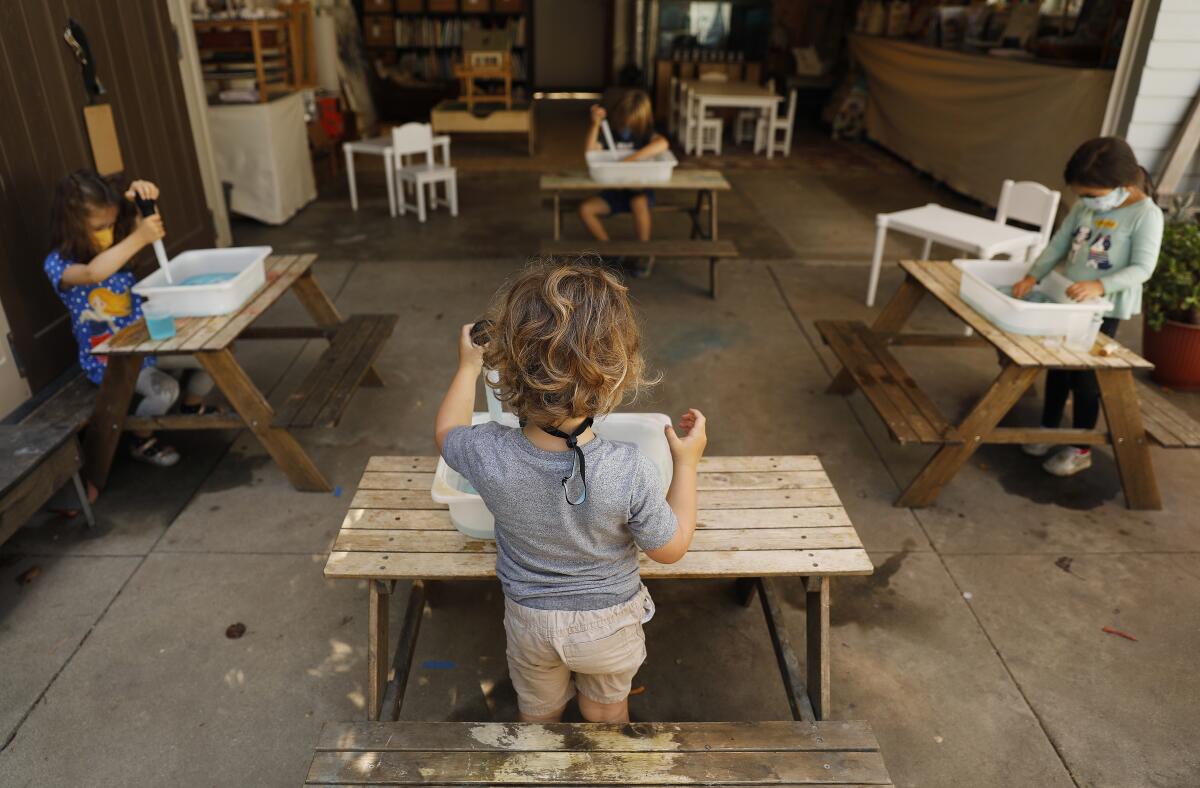 Children with masks play at separate wooden tables on a patio at Voyages Preschool.