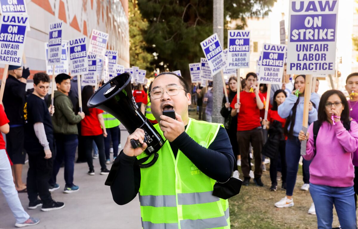 A man wearing a reflective vest uses a megaphone as he walks between rows of protesters with signs.