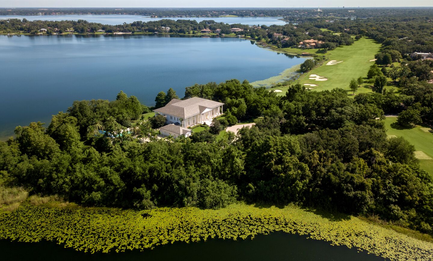 An aerial view of the waterfront home on a peninsula and alongside the greens and sand traps of a golf course.