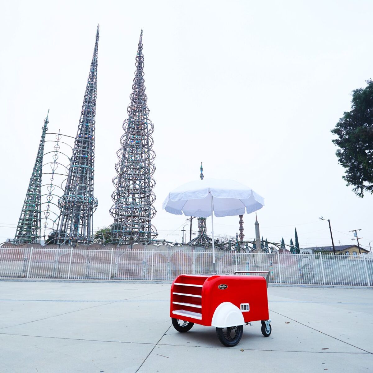 A cherry red sidewalk vending cart with a white umbrella is shown parked in front of the Watts Towers