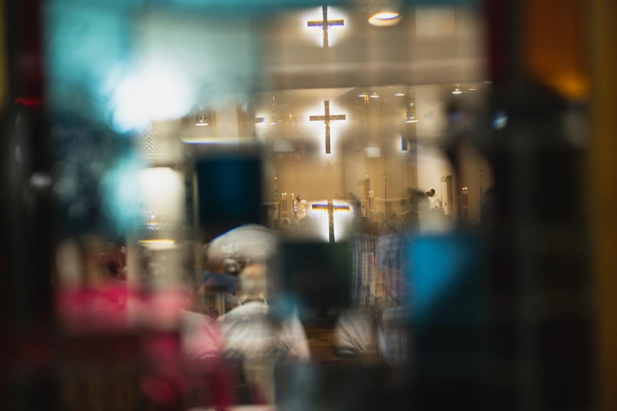 A cross is seen on a wall of a church, with people seated in pews.