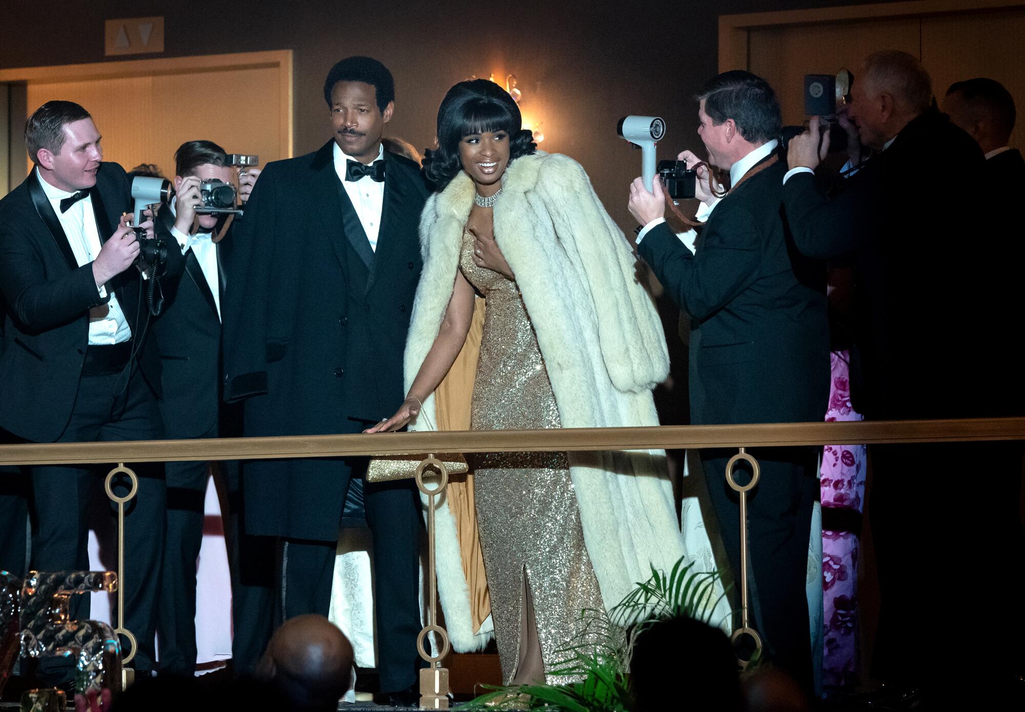 A man in a tux stands next to a woman in a sparkly gold dress and a white fur coat. 