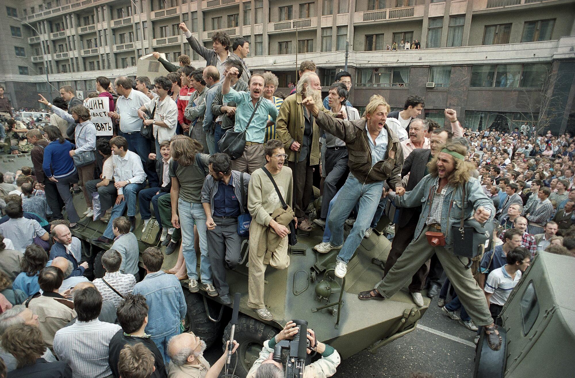 People stand atop a personnel carrier on a street, surrounded by crowds of people in front of a building