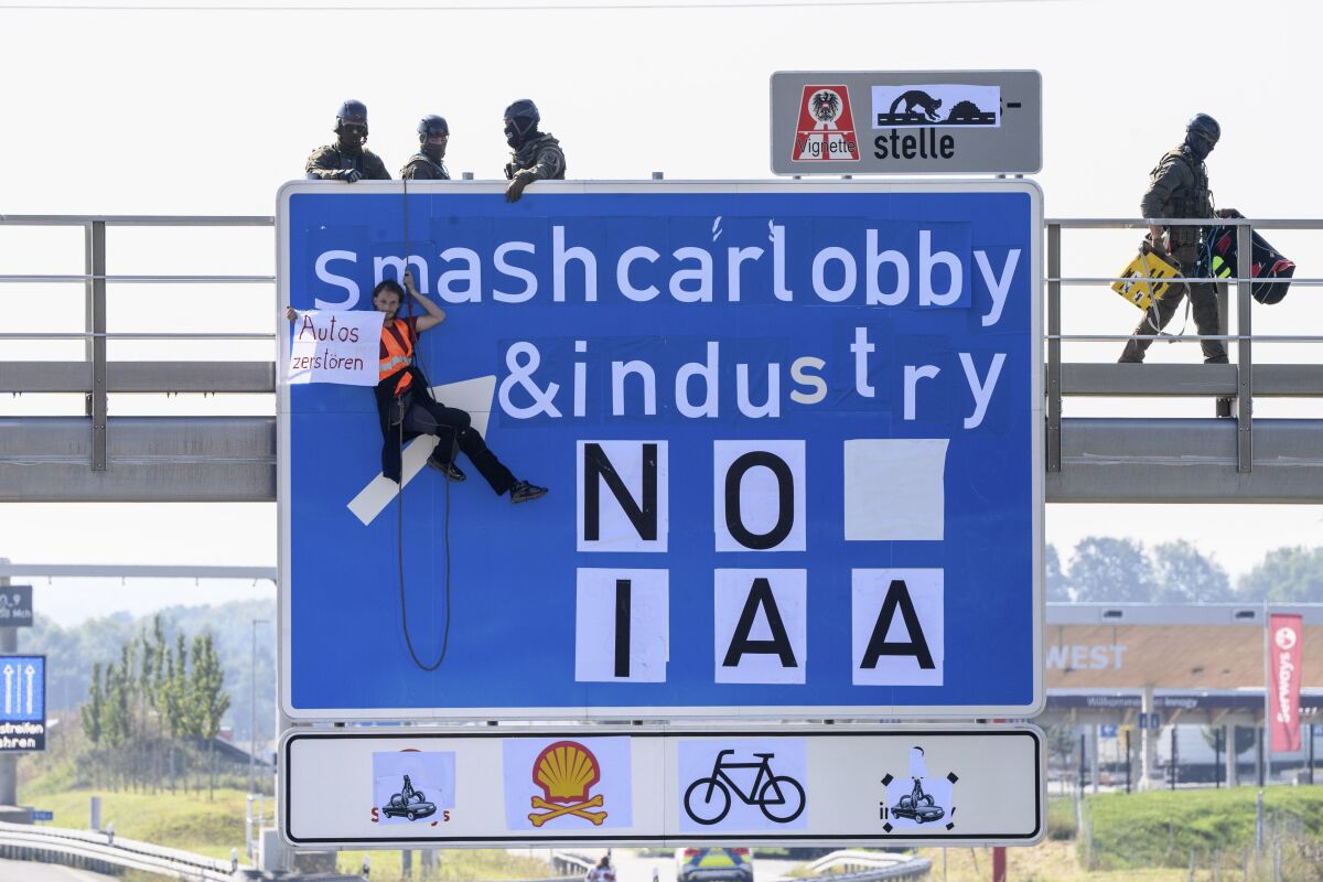 An activist hangs from a gantry over the A9 motorway near Fürholzen in the direction of Munich during a banner campaign, holding a banner with the words "Destroy cars" in his hands while police officers from a special task force get into position on the gantry, Tuesday, Sept.7, 2021. The activists have pasted over the traffic sign with the words "smashcarlobby & industry - NO IAA". (Matthias Balk/dpa via AP)