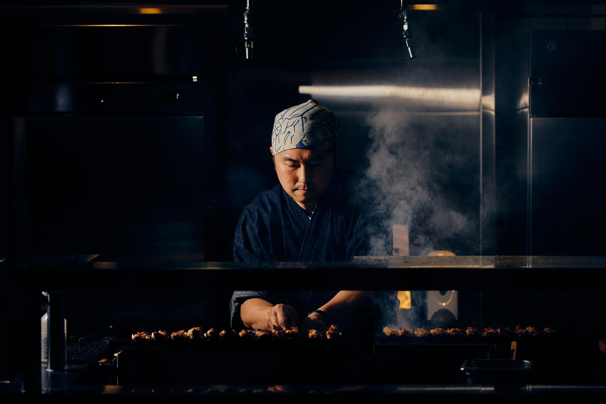 A man faces a line of meat skewers on a smoking grill.