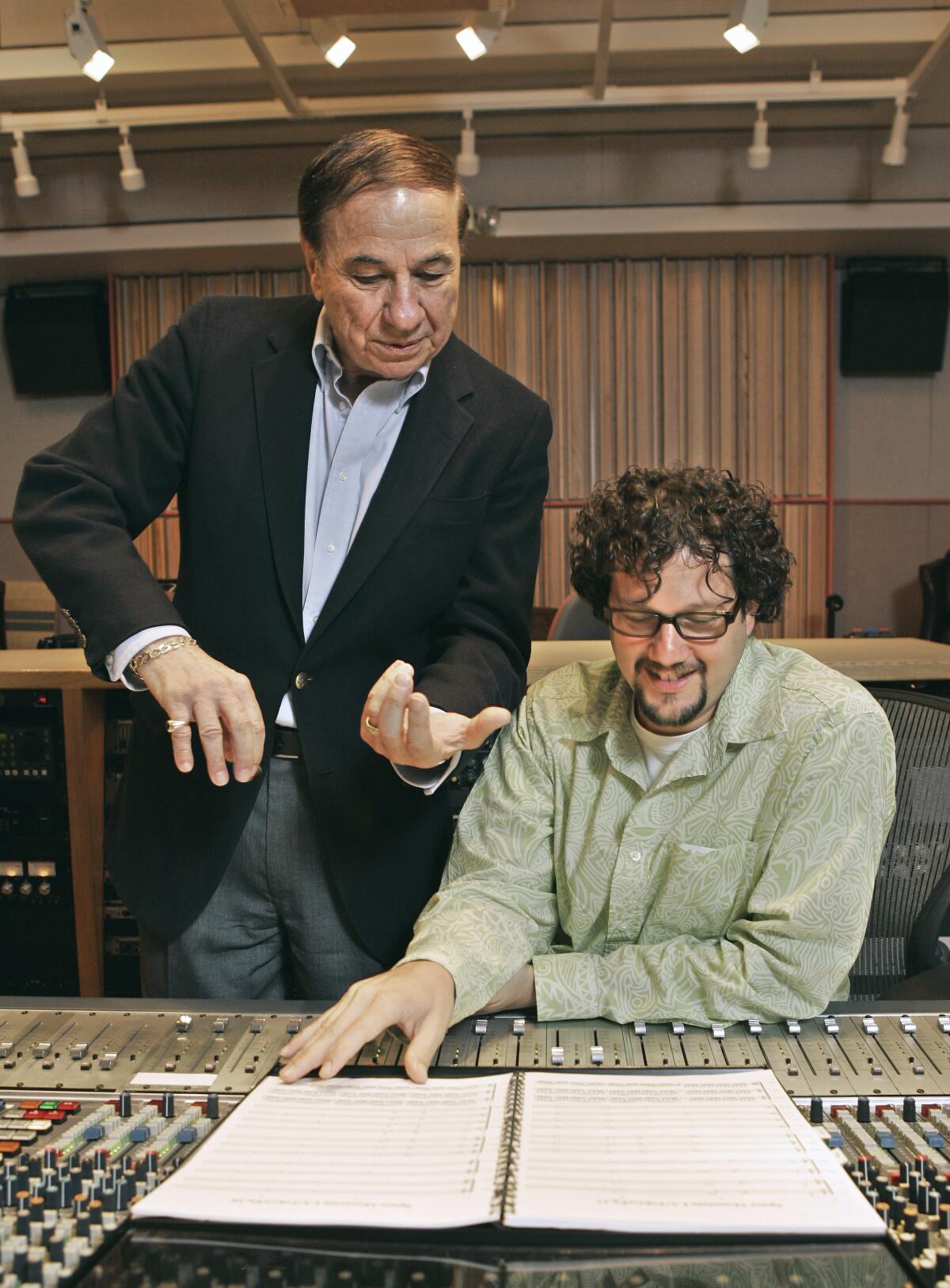 A man in a suit stands next to a seated man as they look at a musical score in a recording studio