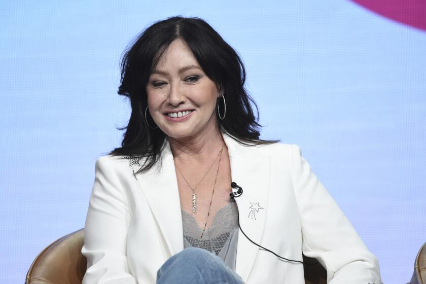 Shannon Doherty in a white blazer, jeans and a grey shirt sitting on a chair with her legs crossed