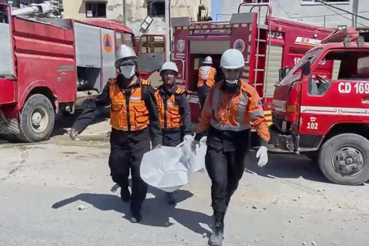 Emergency workers in hard hats and orange vests carry a white body bag near red fire trucks