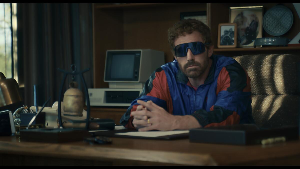 Ben Affleck in a blue and red windbreaker and big sunglasses sitting behind a desk with his hands clasped