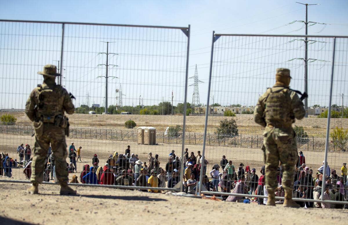 ARCHIVO - Varios migrantes esperan junto a la cerca fronteriza ante la mirada de guardias nacionales 