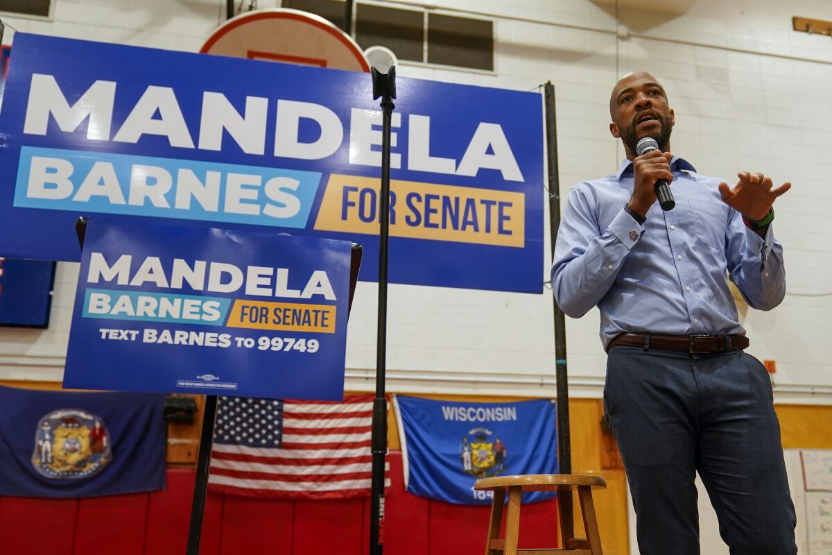 A man with a dark beard holds a microphone as he speaks near a sign that reads, "Mandela Barnes for Senate"