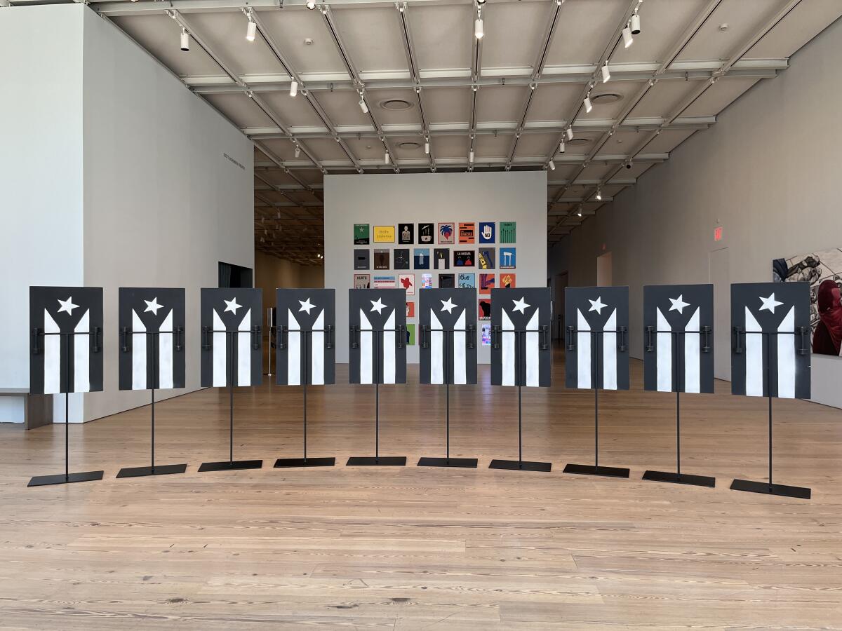 A row of shields painted with a black and white version of a Puerto Rican flag stand on metal poles in a gallery