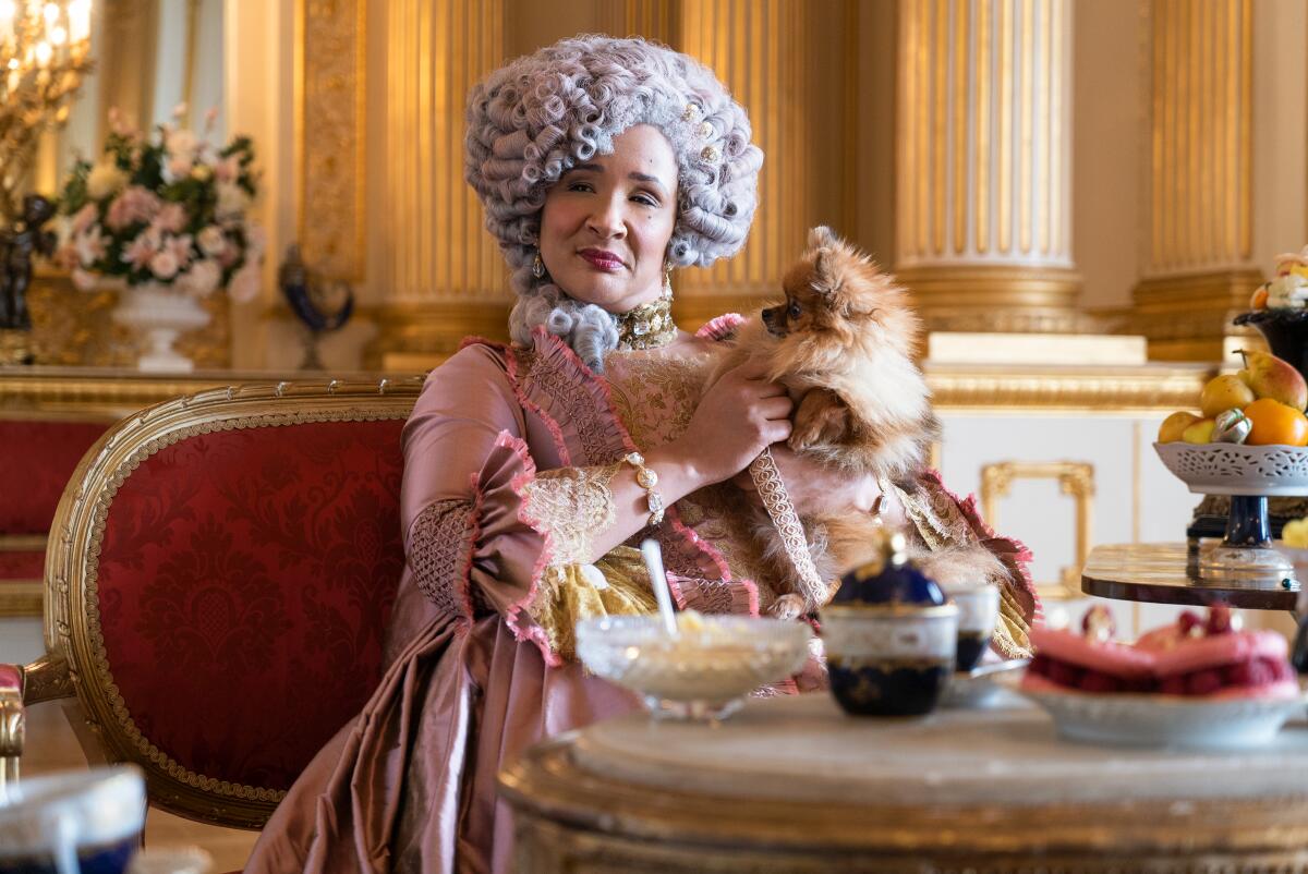 A woman holds a small dog while wearing a tall wig and period gown