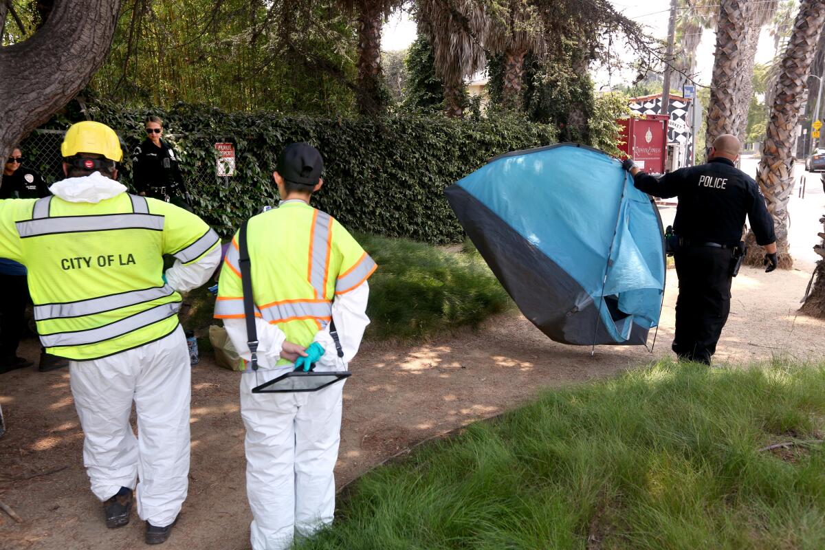 Two people in reflective vests stand on a sidewalk as a police officer carries a small tent away.