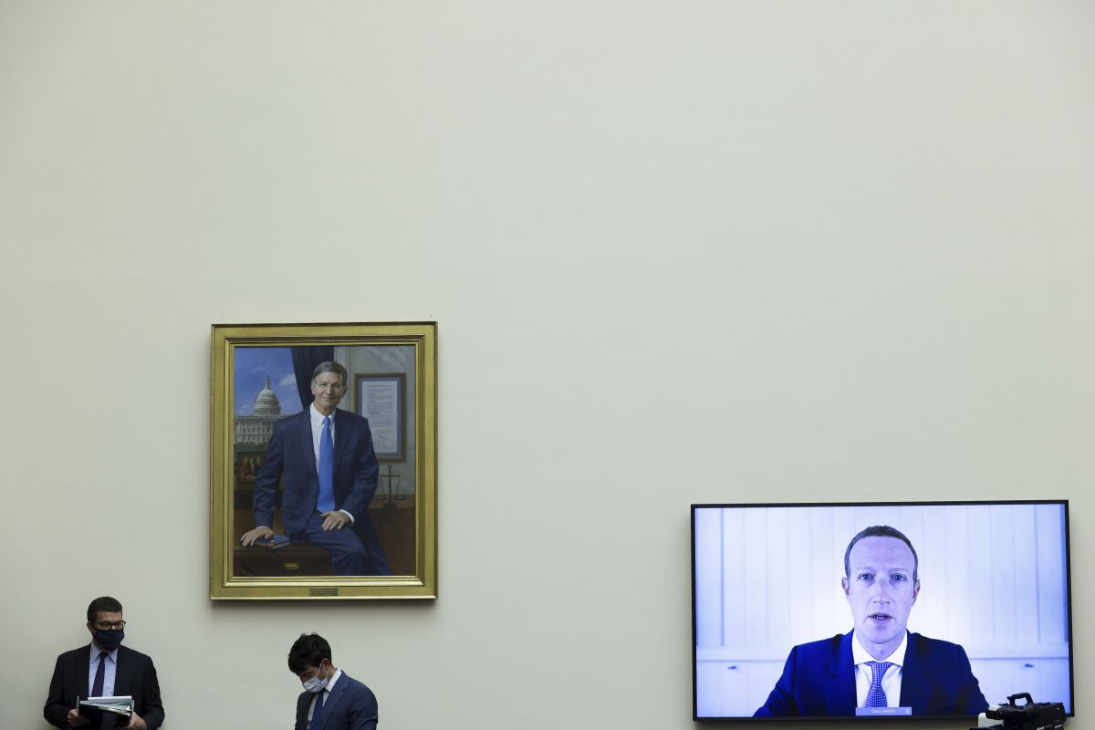 Mark Zuckerberg is shown testifying via video link before Congress next to a painting as workers in masks mill about.