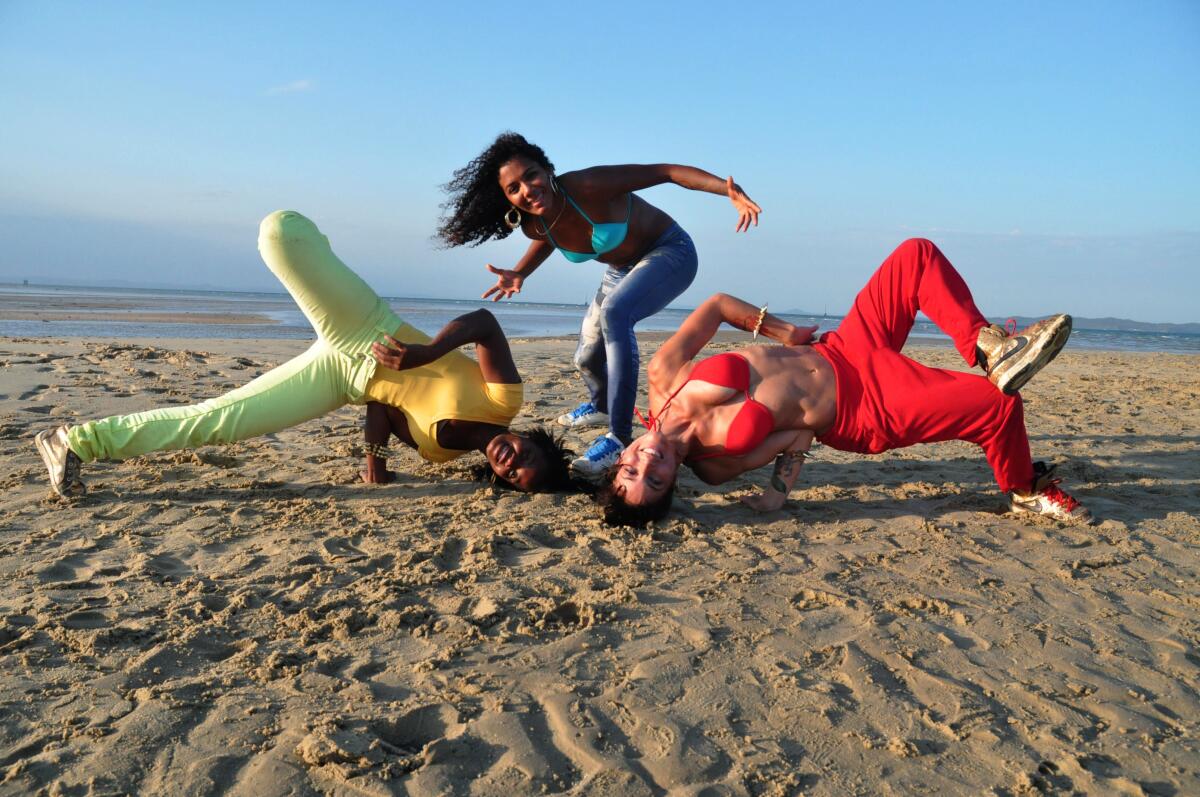 Three people dance on the beach in bright clothing.