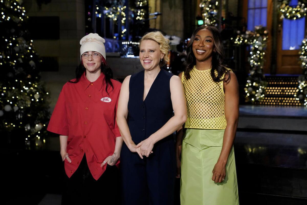 Billie Eilish in a red shirt, Kate McKinnon in a black top, and Ego Nwodim in a yellow top and green skirt on a stage.