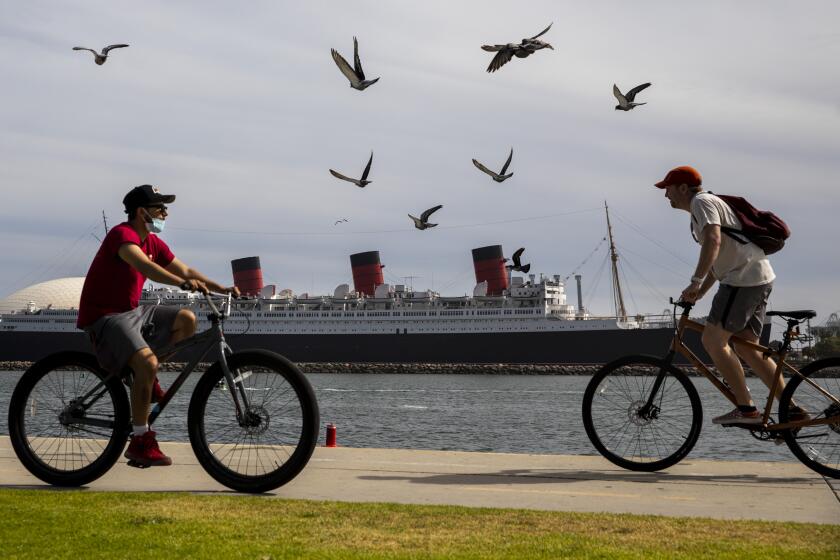 Long Beach, CA - May 25: Bkera and pigeons in Shoreline Aquatic Park are seen in the foreground, with the Queen Mary ship in the distance, docked in Long Beach, CA, photographed Tuesday, May 25, 2021. The ship has been a tourist destination and hotel for years and is now in danger of capsizing according to a recent inspection report. (Jay L. Clendenin / Los Angeles Times)