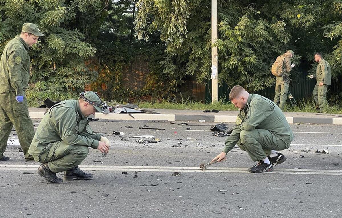Russian investigators at the scene of a car explosion on a street with debris near trees.